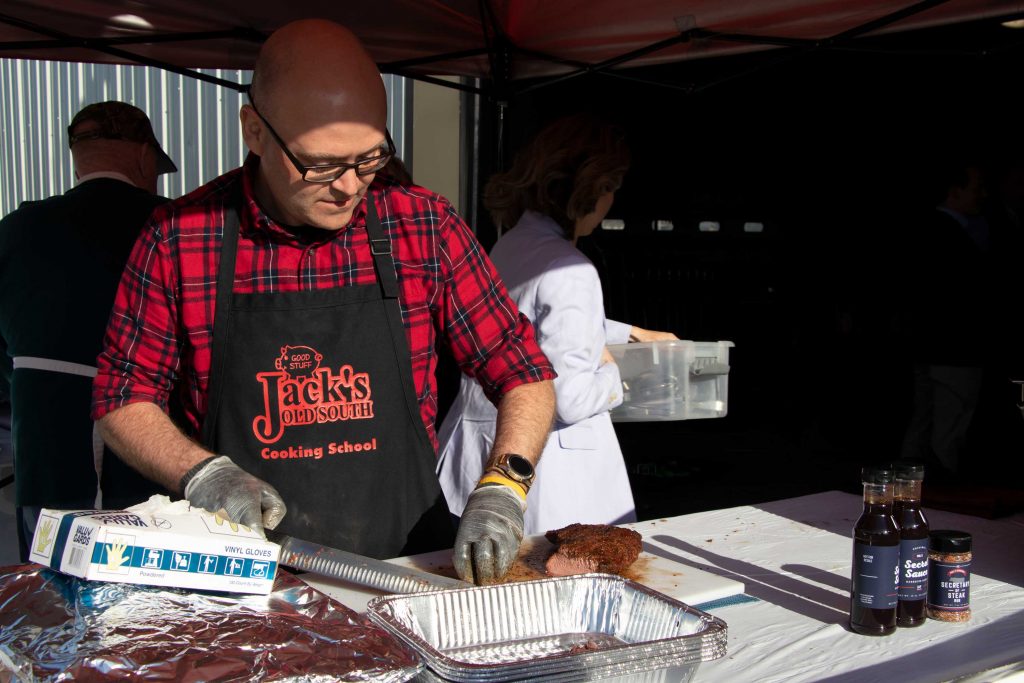 A balding man in a red plaid shirt and cooking apron slices some barbecue meat on a cluttered table